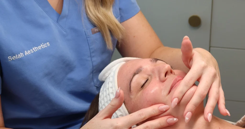 A Selah Aesthetics aesthetician in blue scrubs gives a facial massage during Skin Peel Treatments in Fairfield, AU, in a modern clinic with a marble backsplash.